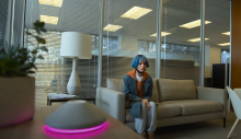 Screenshot from KIMI.  Female sits on a couch in an office, looking at a virtual assistant device on the table. 